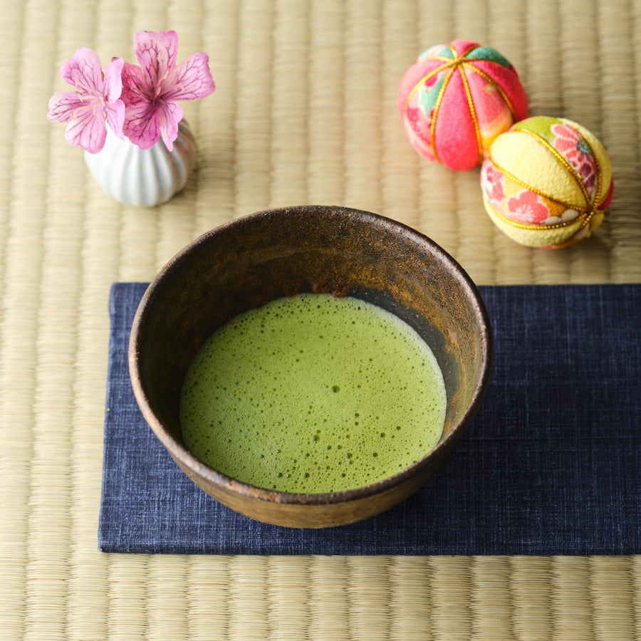 Whisked organic ceremonial grade matcha in a rustic dark raku chawan on an indigo cloth and tatami mat, with pink flowers and temari balls