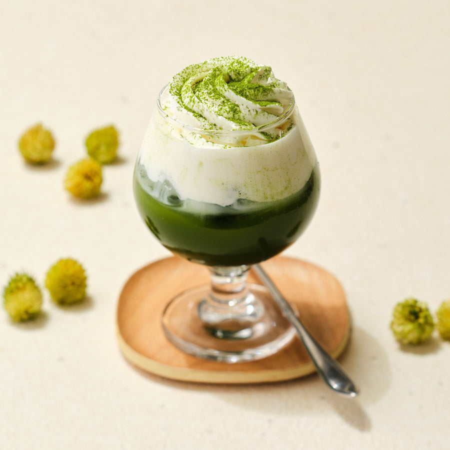 Layered iced matcha with cream and a dusting of matcha in a stemmed glass on a wooden coaster