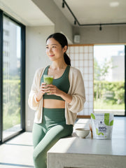 Woman drinking iced matcha in a bright room, with a 100% Organic Japanese Matcha 50 g pouch, chawan and whisk on the counter beside her.