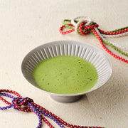 Bowl of whisked matcha in a fluted gray bowl with braided silk cords on a textured background