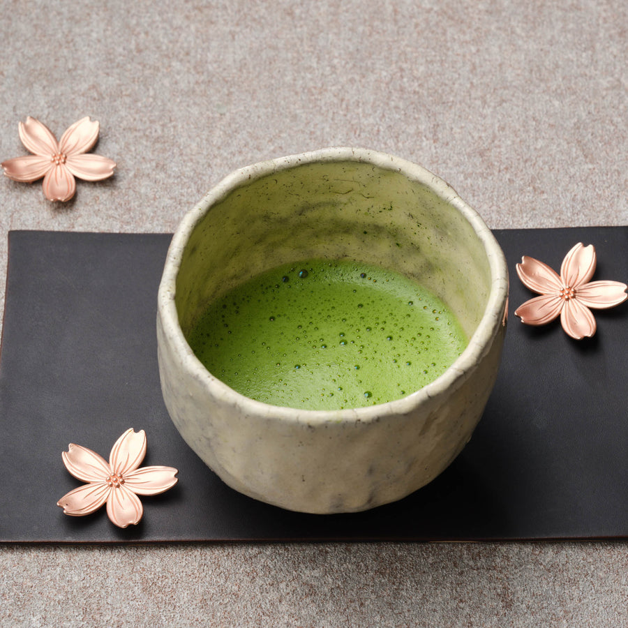 Whisked organic Okumidori matcha in a rustic cream raku chawan on a black tray, with copper cherry blossom ornaments