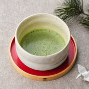 Whisked organic Saemidori matcha in a white glazed chawan on a red lacquer tray, with a pine sprig and a small porcelain crane