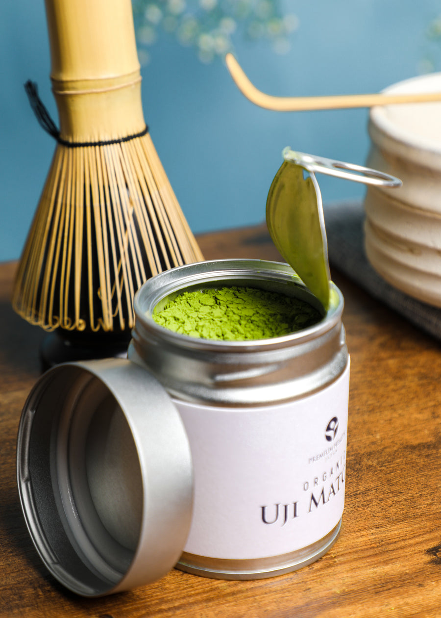 Opened tin of Organic Uji Matcha 50g showing vibrant green powder beside a bamboo whisk and chawan, representing authentic Kyoto tea craftsmanship.