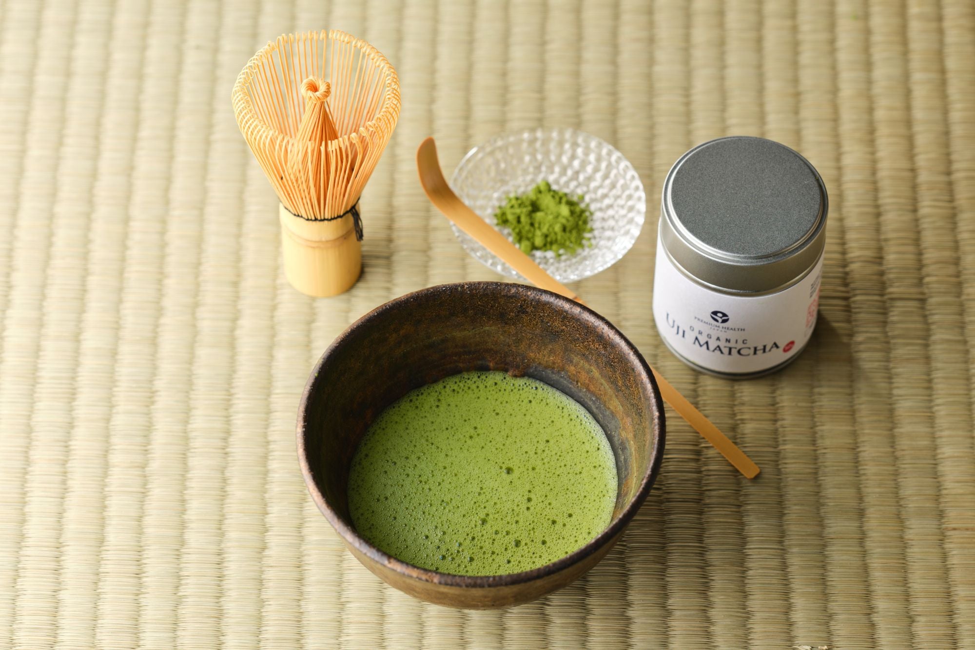 Organic Uji matcha 50g silver tin with a whisked bowl of matcha, bamboo chasen and chashaku, and a glass dish of green powder on a tatami mat