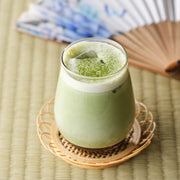 Iced matcha latte made with organic ceremonial grade Uji matcha in a glass on a bamboo coaster, with a Japanese folding fan behind