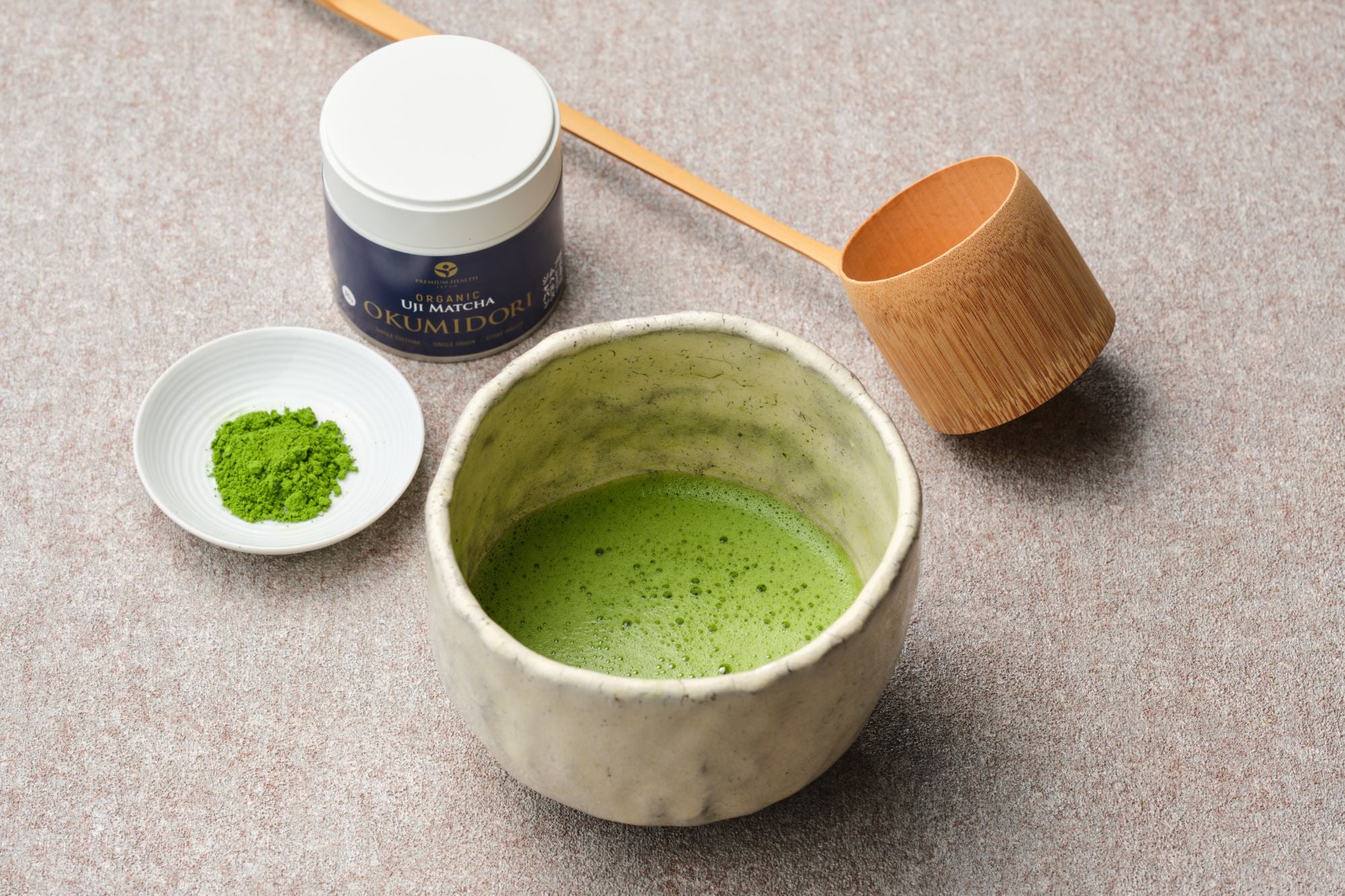 Organic Uji matcha Okumidori 30g navy tin with a whisked bowl of matcha, bamboo ladle and a dish of green powder on a stone surface