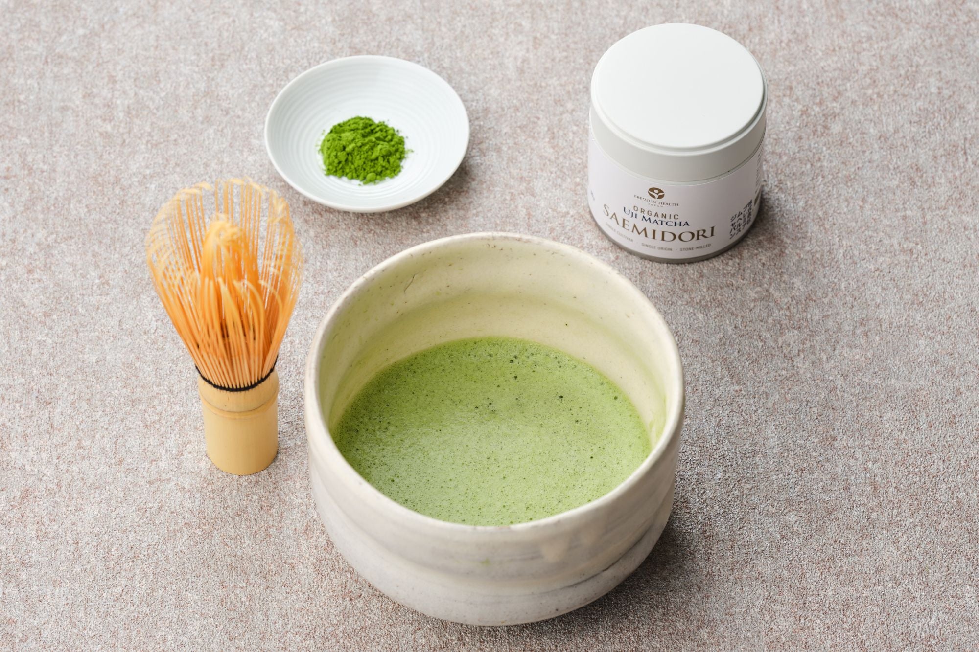 Organic Uji matcha Saemidori 30g white tin beside a whisked bowl of matcha, bamboo chasen and a dish of green powder on a stone surface