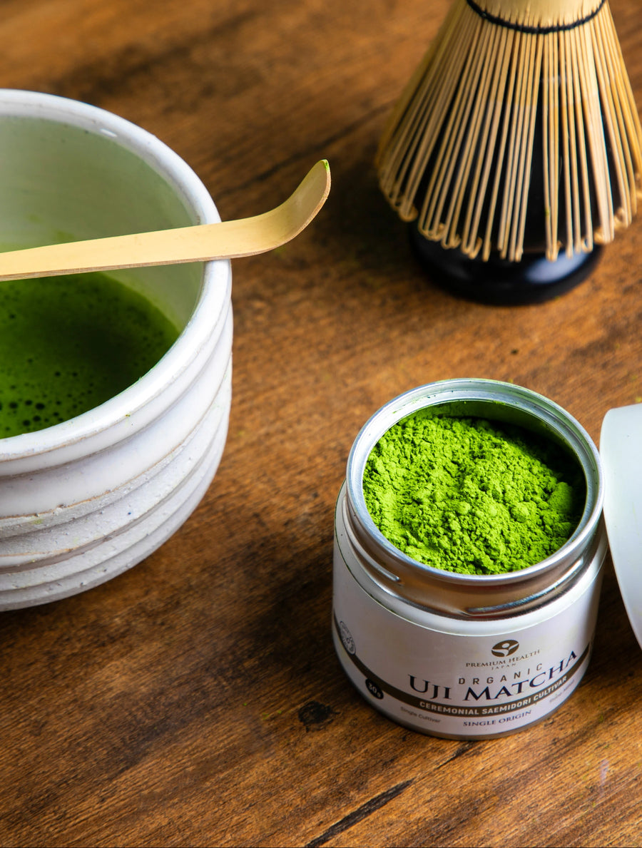Open can of Organic Uji Matcha Saemidori cultivar with vibrant green powder beside a matcha bowl and bamboo whisk on a wooden table.