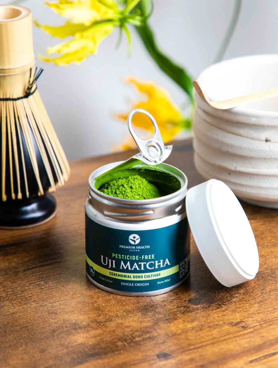 Open can of Pesticide-Free Uji Matcha (Gokō cultivar) showing vibrant green powder beside whisk and chawan on wooden table.
