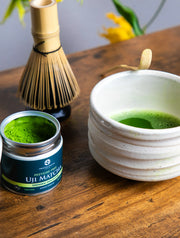 Open jar of pesticide-free Uji Matcha Gokō cultivar beside a whisk and frothy matcha in a white chawan on a wooden table