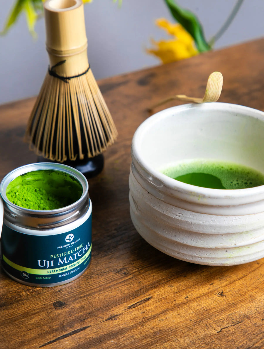 Open jar of pesticide-free Uji Matcha Gokō cultivar beside a whisk and frothy matcha in a white chawan on a wooden table
