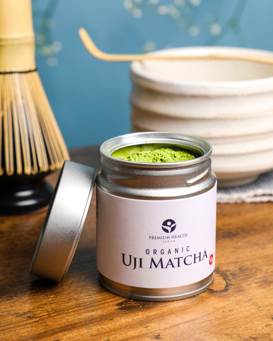 Premium Health Japan Organic Uji Matcha 50g tin with lid off, showing vibrant green tea powder beside a bamboo whisk and chawan in a Kyoto-inspired setting.