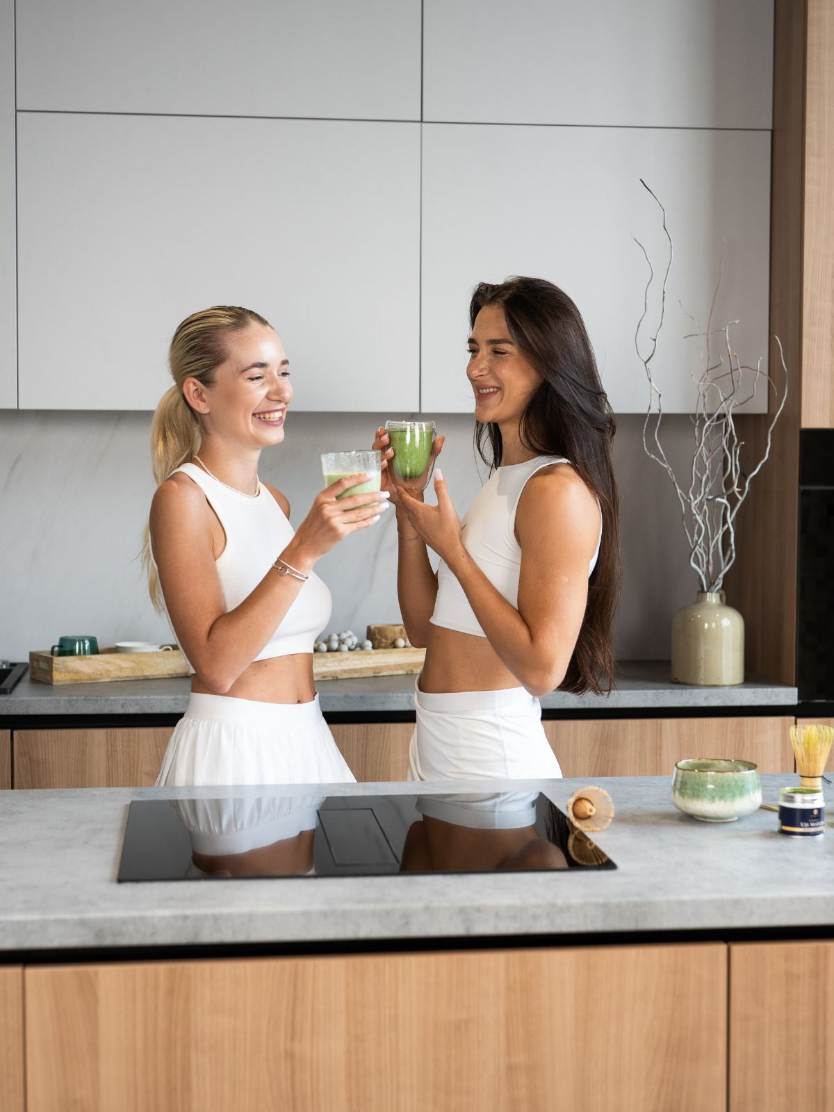 Two women enjoying iced matcha lattes in a modern kitchen with Premium Health Japan Organic Uji Matcha tin visible on the counter.
