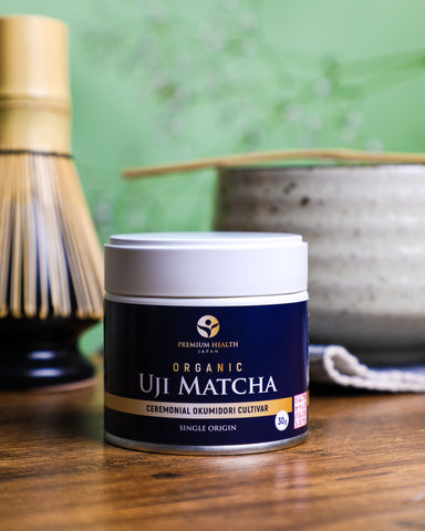 Premium Health Japan Organic Uji Matcha Okumidori cultivar 30g tin beside bamboo whisk and chawan, representing single-origin ceremonial Kyoto matcha.