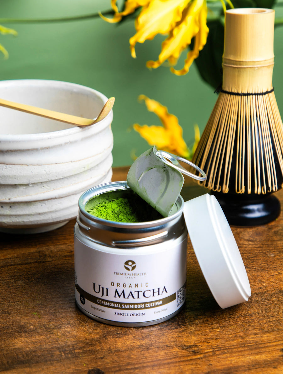 Open can of Organic Uji Matcha Saemidori cultivar with vibrant green powder, whisk and bowl in background on wooden table.