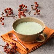 Hot matcha latte made with organic single-cultivar Saemidori Uji matcha in a brown stoneware cup on an orange cloth, with red autumn berries