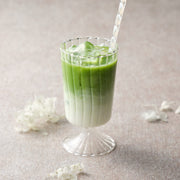 Iced matcha latte made with JAS organic Saemidori matcha, green matcha layered over milk and ice in a fluted stemmed glass with a striped straw and white hydrangea petals