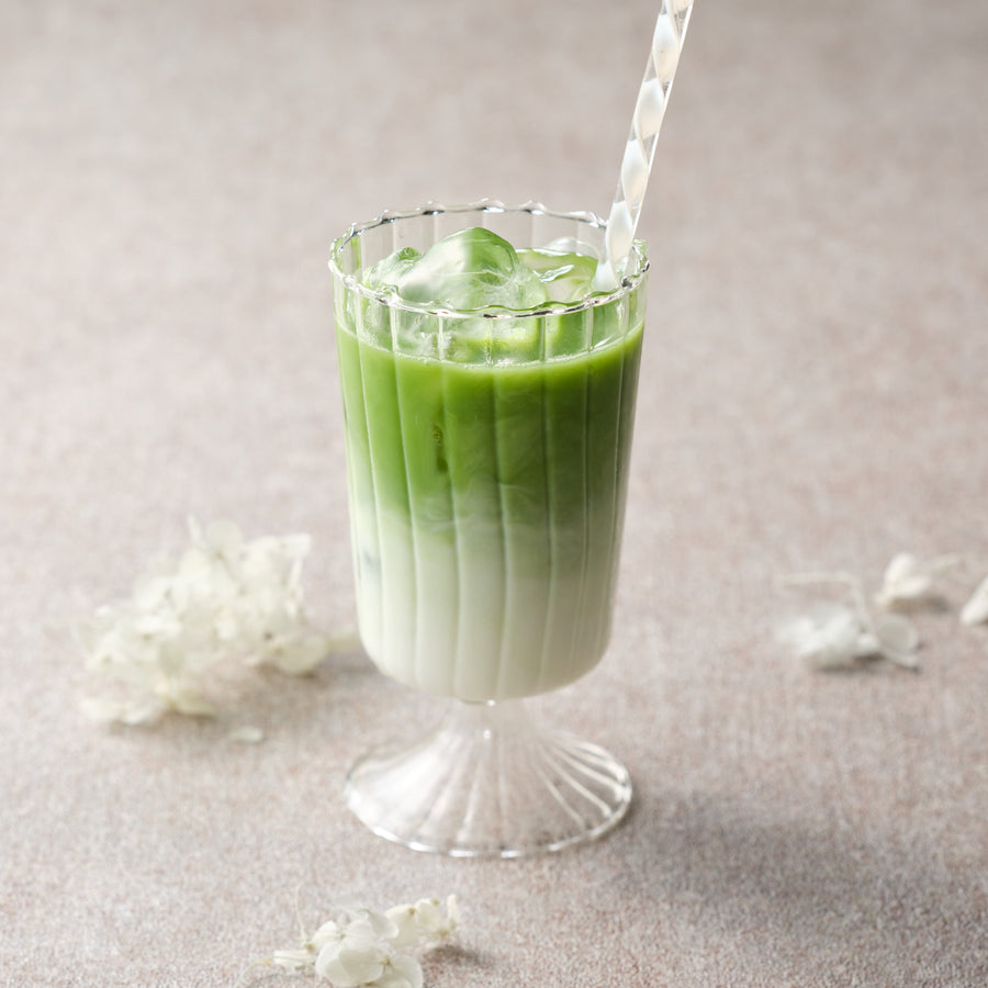 Iced matcha latte made with JAS organic Saemidori matcha, green matcha layered over milk and ice in a fluted stemmed glass with a striped straw and white hydrangea petals