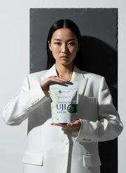 A model in a white suit holding a 100 g pouch of Ceremonial Grade Uji Matcha from Premium Health Japan against a minimalist studio backdrop.
