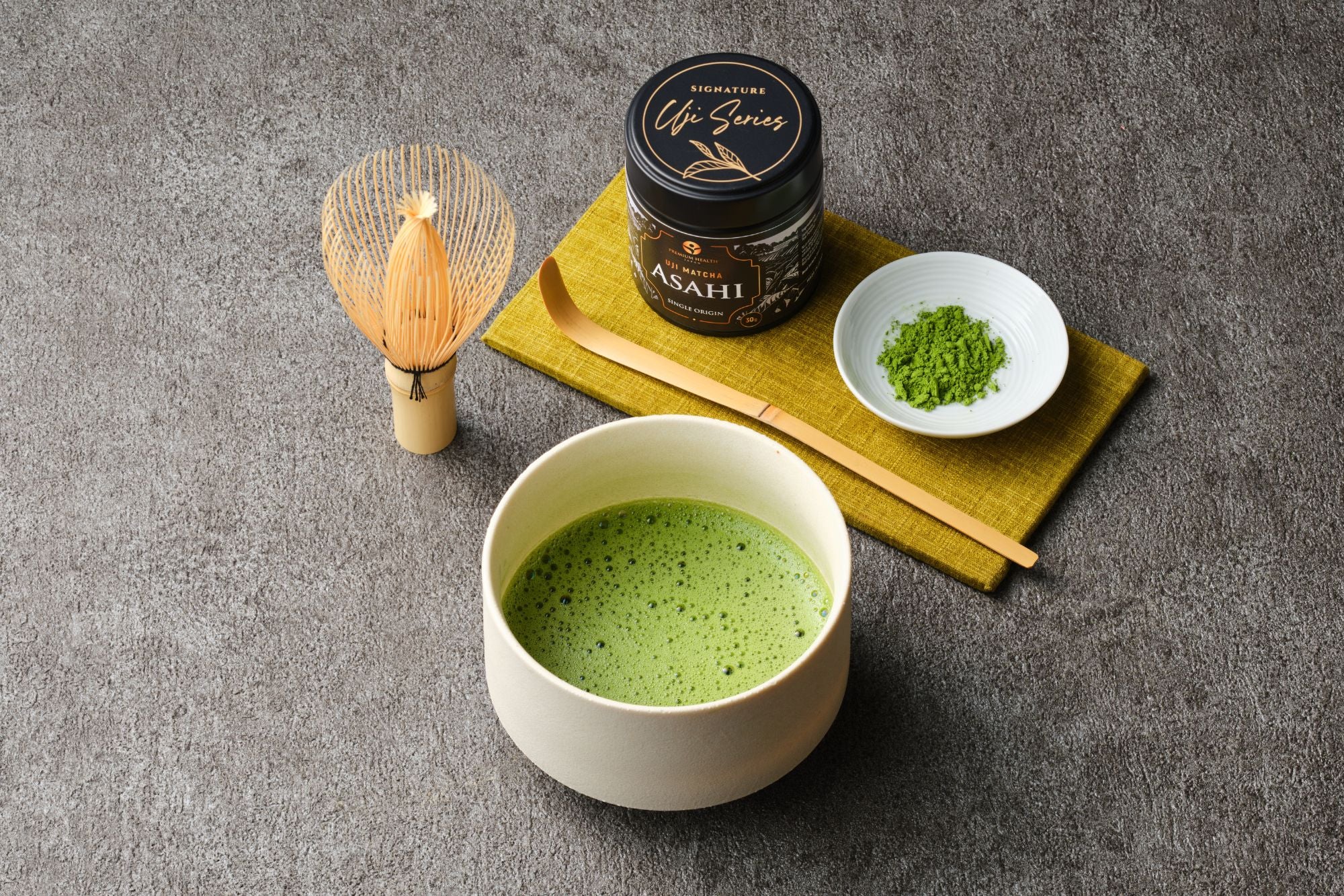 Uji matcha Asahi Signature Uji Series black tin on an olive cloth with a whisked bowl of matcha, bamboo chasen and chashaku, and a dish of green powder on gray stone