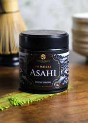 Uji Matcha Asahi single-origin 30g tin on a wooden surface with matcha powder and bamboo whisk in background, highlighting authentic Kyoto craftsmanship.