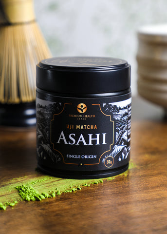 Uji Matcha Asahi single-origin 30g tin on a wooden surface with matcha powder and bamboo whisk in background, highlighting authentic Kyoto craftsmanship.