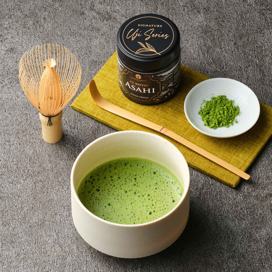 Uji Matcha Asahi single origin 30g tin with bamboo whisk, chashaku scoop, matcha powder and a whisked bowl of matcha on a mustard linen mat