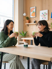 Two people enjoying hot matcha drinks in a café with a tin of Uji Matcha Gokō placed on the table.