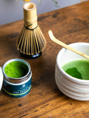 Open tin of Uji Matcha Gokō cultivar beside a whisk and bowl of freshly prepared matcha on a wooden table