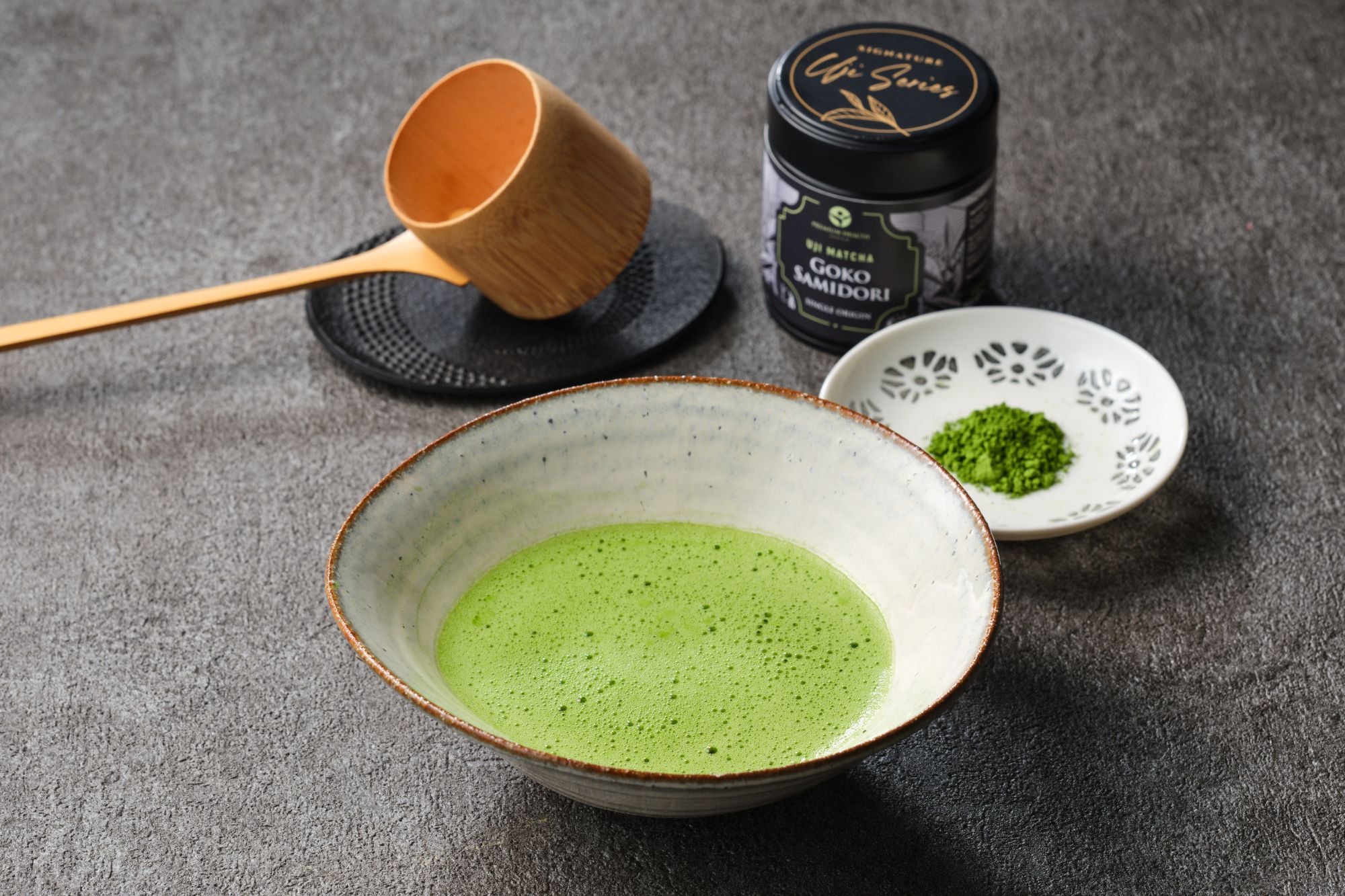 Uji matcha Goko Samidori Signature Uji Series black tin with a whisked bowl of matcha, bamboo ladle on a cast-iron trivet and a dish of green powder on dark stone