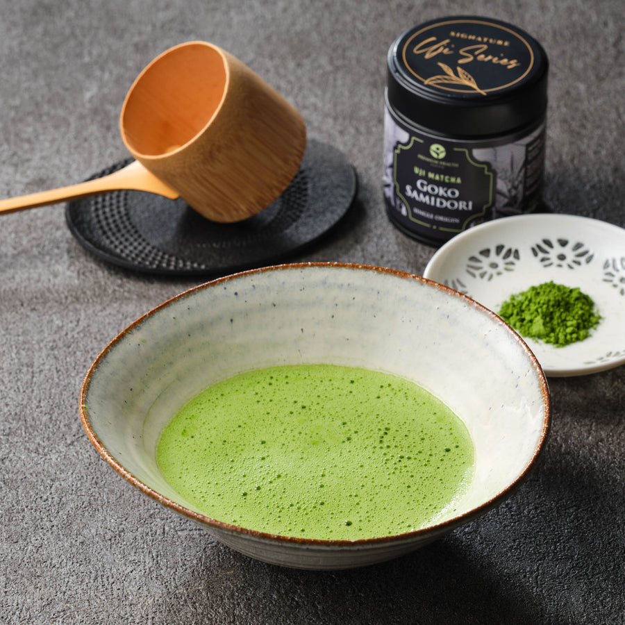 Uji Matcha Gokō × Samidori hand-picked ceremonial blend 30g tin beside a whisked bowl of matcha, bamboo hishaku ladle on a cast-iron trivet and matcha powder on a white dish