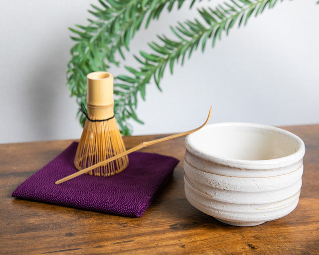 Traditional Japanese bamboo whisk and chashaku beside a white ceramic matcha bowl on a wooden table with green foliage background.