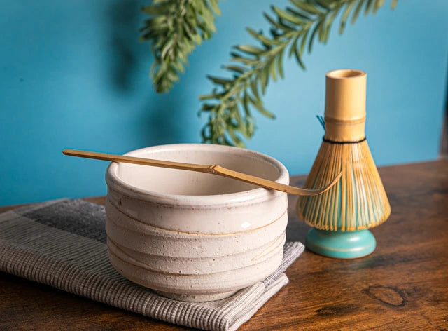 White ceramic chawan with bamboo chashaku across the rim; bamboo whisk on turquoise stand (kusenaoshi) against a blue backdrop.
