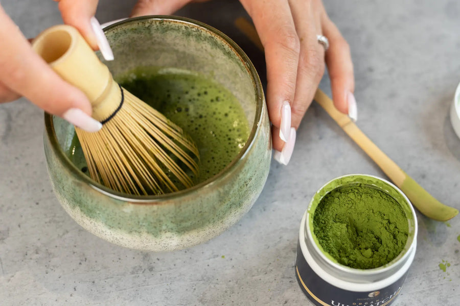 Hand whisking organic Uji matcha in a green chawan with bamboo whisk, beside open matcha tin and chashaku, showing authentic Japanese tea preparation.